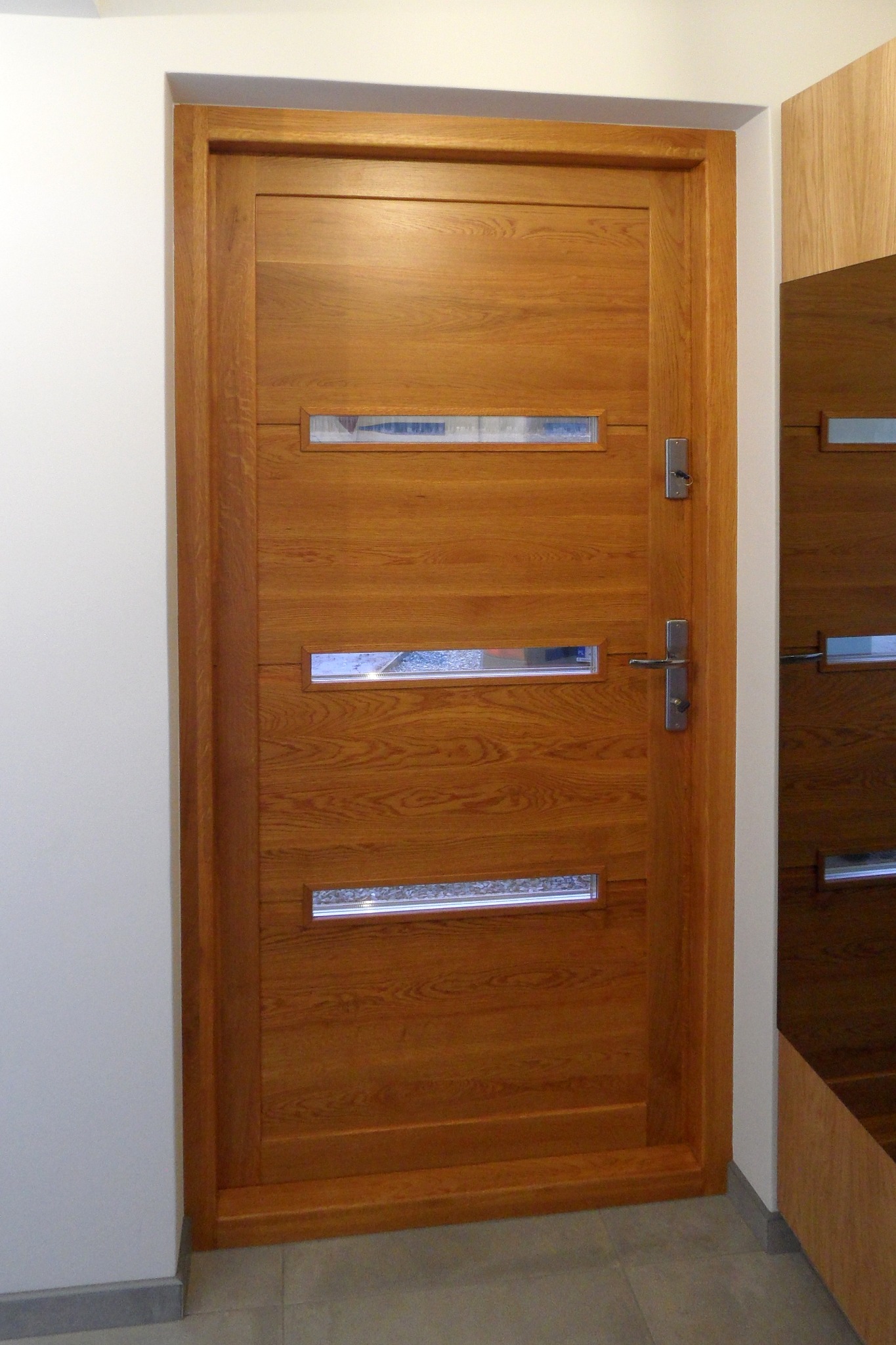 Nowoczesne drzwi wejściowe z drewna o ciepłym odcieniu, z trzema poziomymi przeszkleniami z dekoracyjnym szkłem, widoczny zamek z kluczem i klamka.