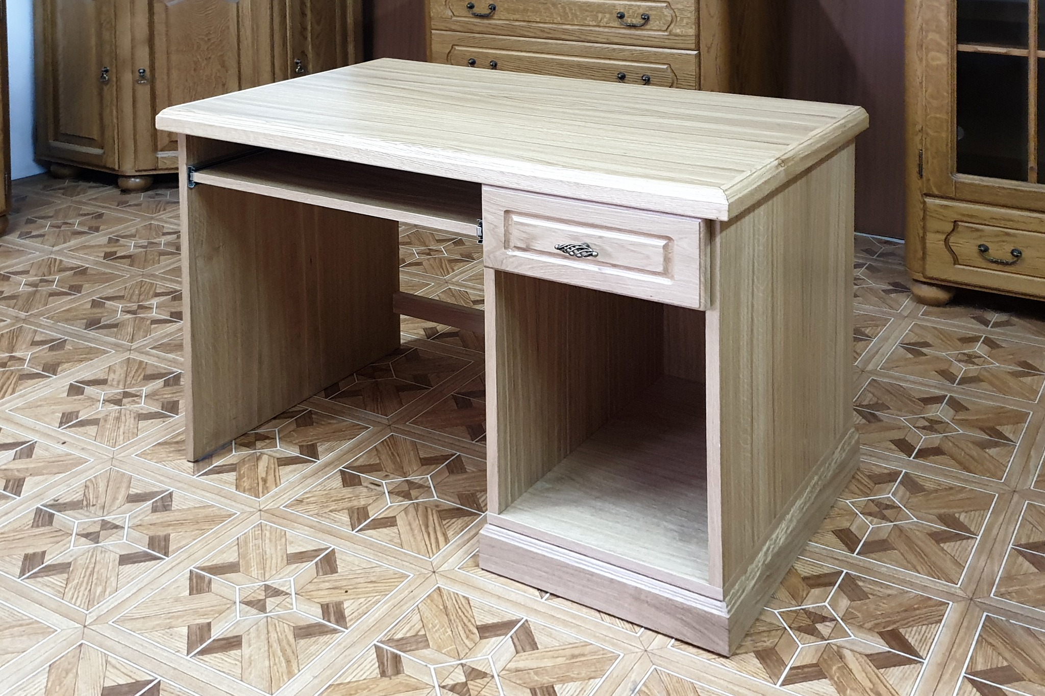 Biurko komputerowe z jasnego drewna z wysuwaną półką na klawiaturę i szufladą z ozdobnym uchwytem, na tle podłogi z wzorzystych płytek ceramicznych i mebli w podobnym stylu.