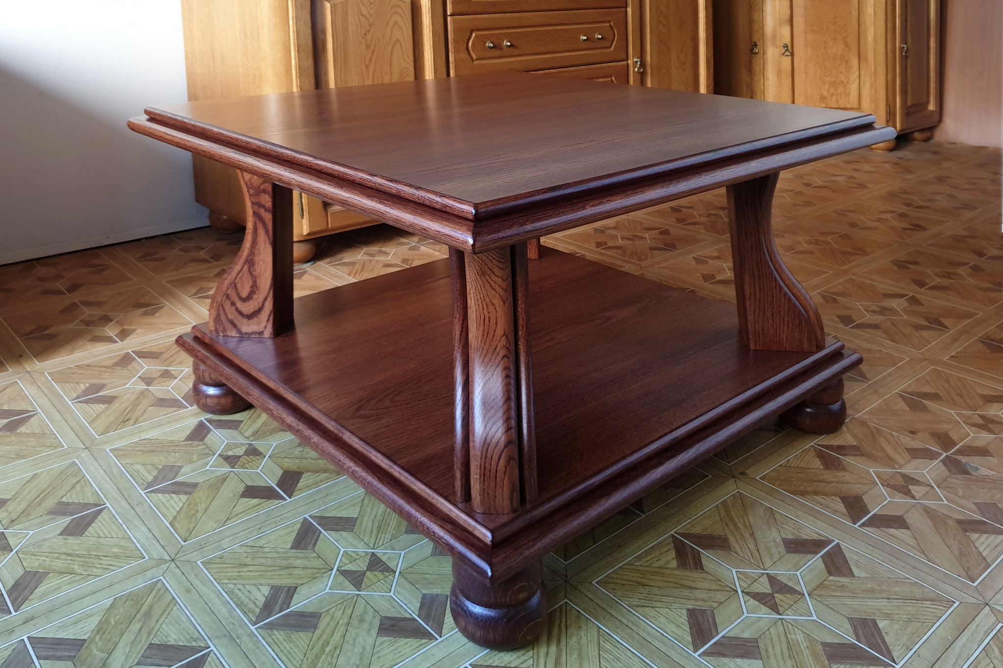 Kwadratowy stolik kawowy z ciemnego drewna z półką, ustawiony na podłodze z geometrycznym wzorem, widoczne również fragmenty drewnianych mebli w tle.