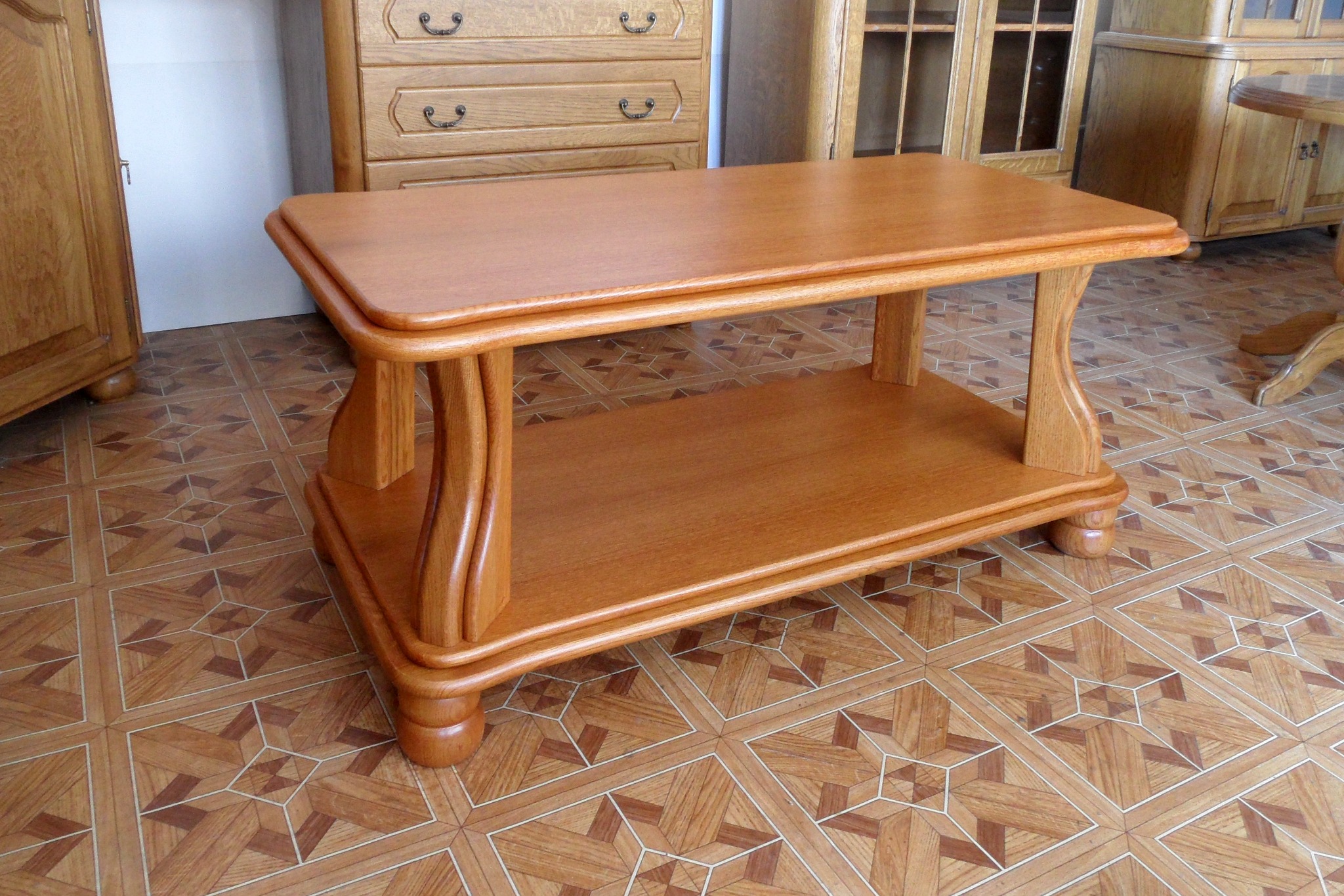 Drewniany stolik kawowy z półką, o zaokrąglonych krawędziach i toczonych nogach, w otoczeniu mebli w podobnym stylu i kolorystyce, na podłodze z wzorzystych paneli.