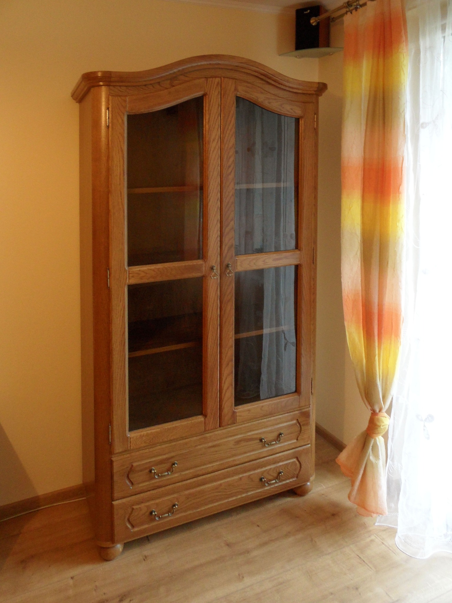 Drewniana witryna z przeszklonymi drzwiami i dwoma szufladami, w klasycznym stylu, na tle beżowej ściany i okna z kolorową zasłoną.