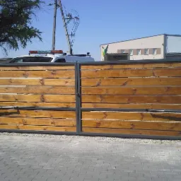 Dwuskrzydłowa brama wjazdowa z drewnianym wypełnieniem i szarą ramą oraz automatyką, widoczna na tle kostki brukowej i budynku.