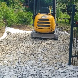 Żółta mini koparka JCB na żwirowej drodze, widoczna brama wjazdowa i przygotowane podłoże z białej geowłókniny, teren zarośnięty zielenią.