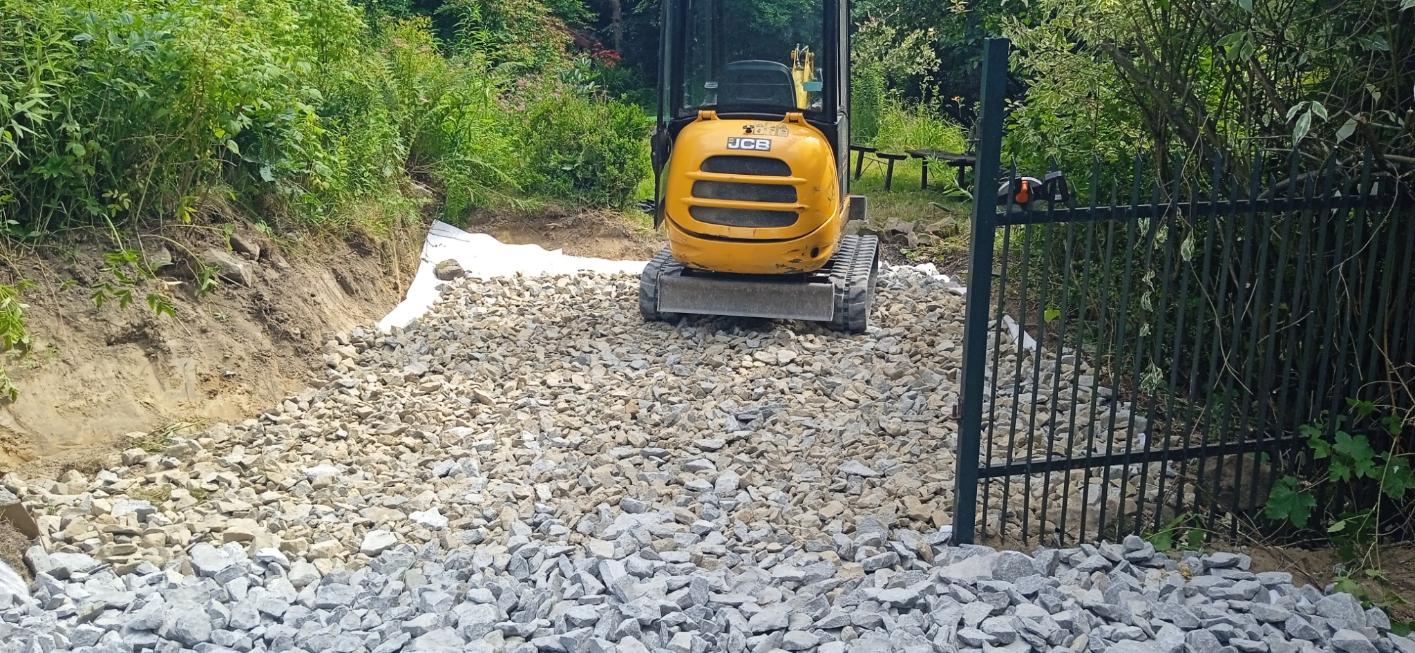 Żółta mini koparka JCB na żwirowej drodze, widoczna brama wjazdowa i przygotowane podłoże z białej geowłókniny, teren zarośnięty zielenią.