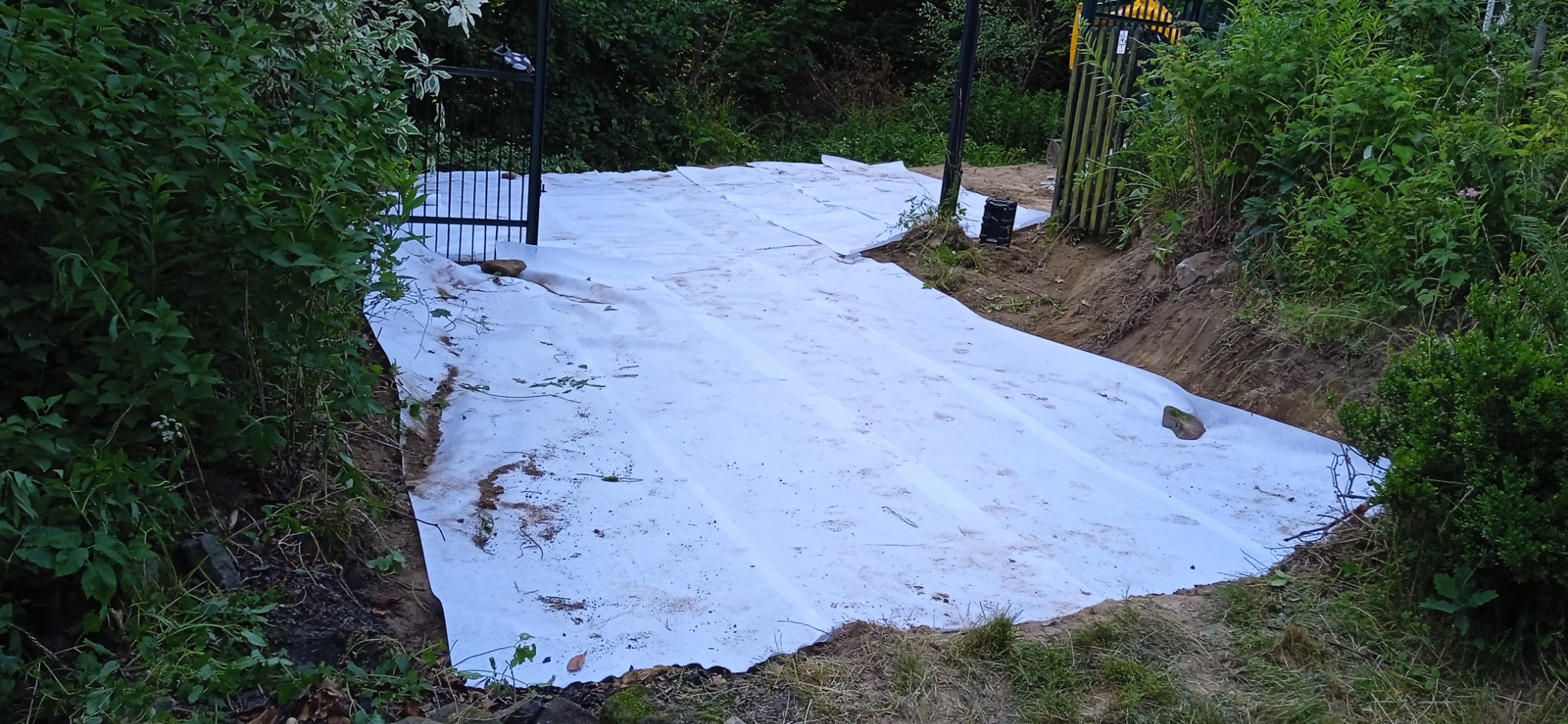 Biała agrowłóknina rozłożona na przygotowanym podłożu, widoczna brama w tle i bujna roślinność po bokach.