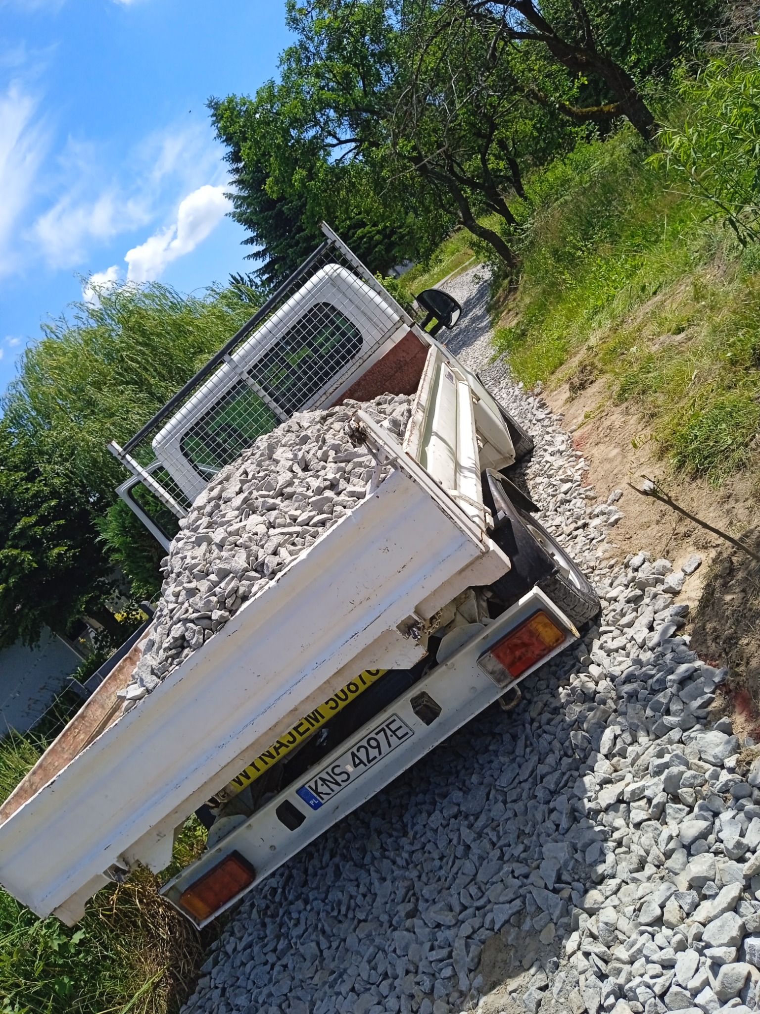 Biały samochód dostawczy z otwartą paką, wypełnioną szarym kruszywem, stojący na drodze wysypanej tym samym materiałem, widok z boku, z zielenią w tle i błękitnym niebem z chmurami.