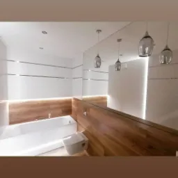 Nowoczesna łazienka z podświetlaną wanną, drewnopodobnymi panelami i lustrem na całej ścianie, wisząca toaleta oraz designerskie lampy zwisające z sufitu.