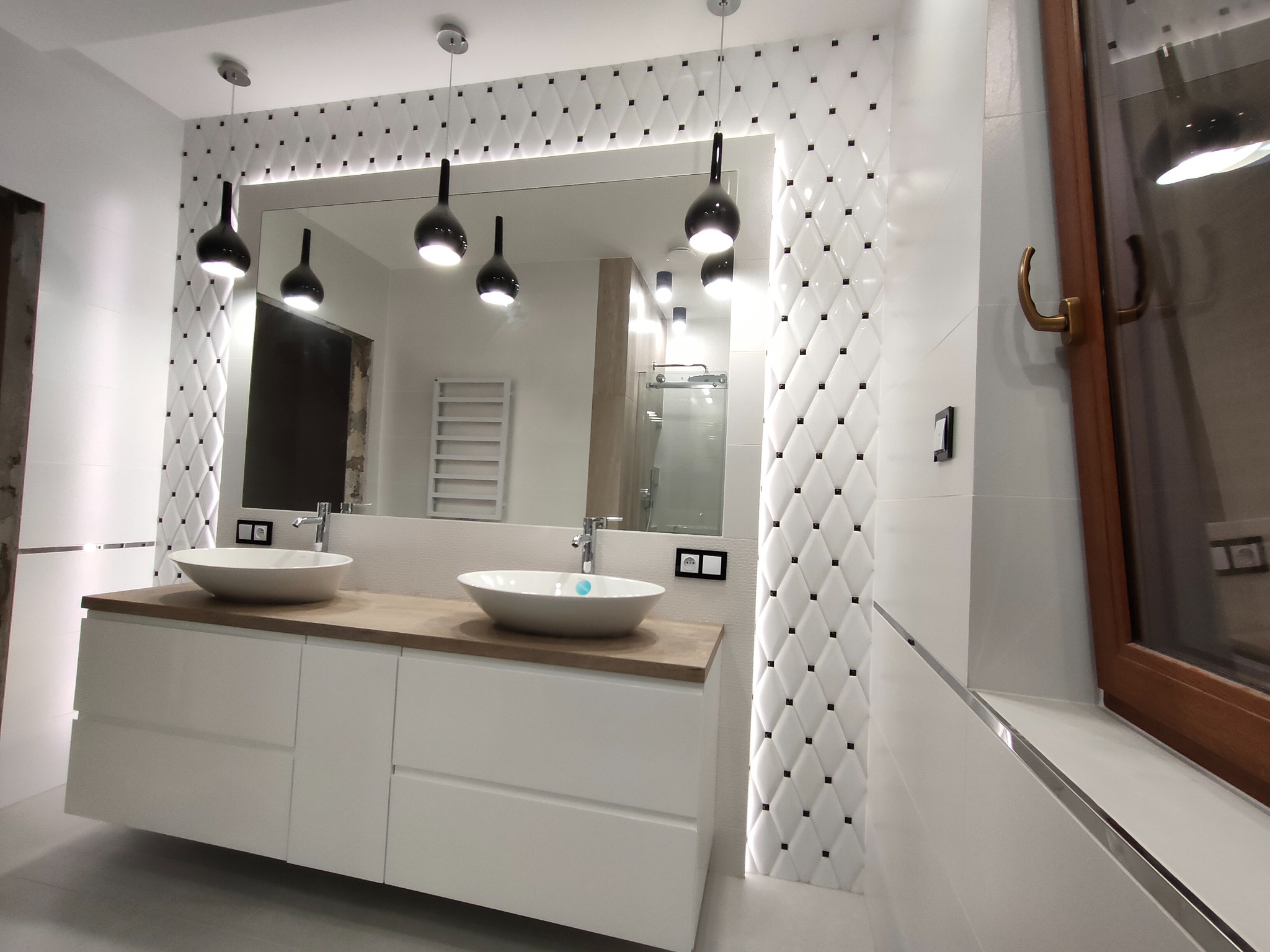 Nowoczesna łazienka z podwójną umywalką nablatową na szafce z białymi frontami, drewnianym blatem, dekoracyjnymi białymi płytkami z czarnymi akcentami i wiszącymi lampami w kształcie kropli.