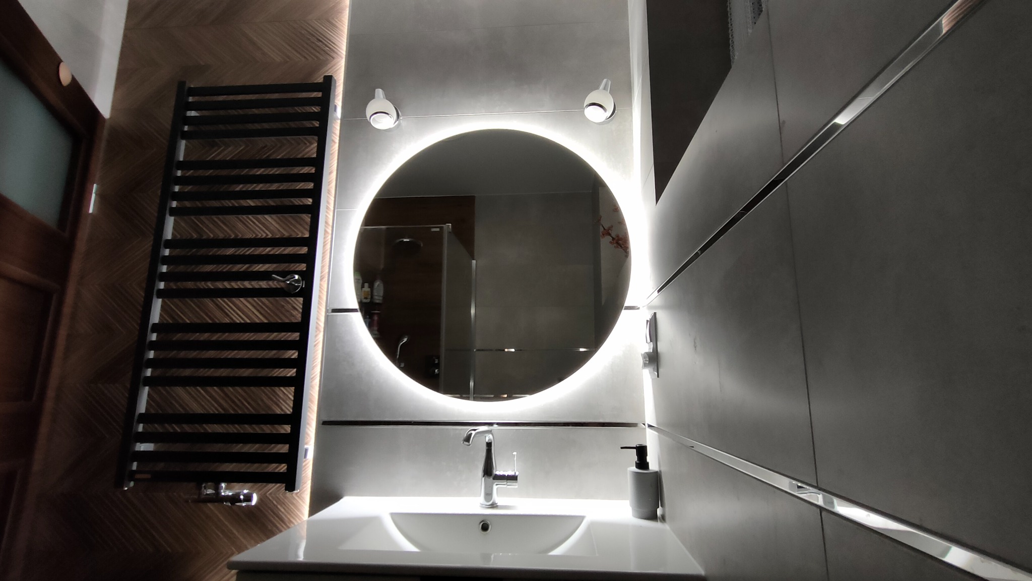 Nowoczesna łazienka z okrągłym lustrem podświetlanym LED, grzejnikiem drabinkowym w kolorze ciemnego brązu, umywalką wpuszczaną w blat i szarą armaturą.