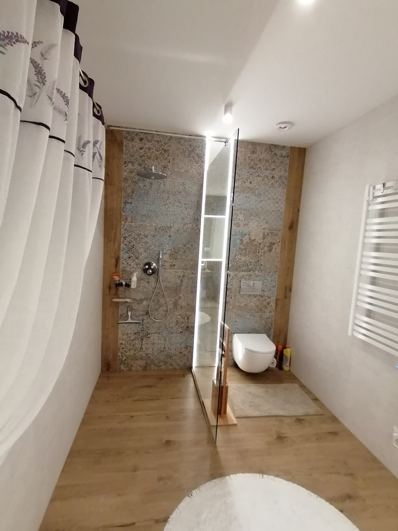 Nowoczesna łazienka z kabiną prysznicową typu walk-in, wykończona płytkami imitującymi kamień, drewnopodobną podłogą i zasłoną w lawendowy wzór.