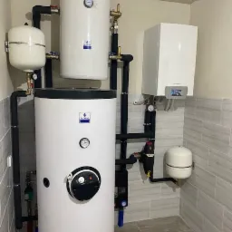 Zestaw urządzeń grzewczych w kotłowni: duży biały zasobnik ciepłej wody z czarnym panelem sterowania, mniejszy zbiornik wyrównawczy, wiszący kocioł gazowy i rury w izolacji, na tle szarych płytek...