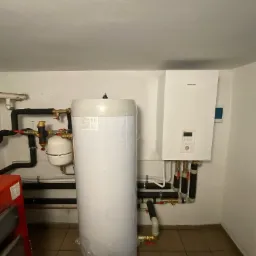 Biały zasobnik ciepłej wody użytkowej, obok wiszący na ścianie biały moduł pompy ciepła Panasonic, widoczne rury i zawory, instalacja w pomieszczeniu technicznym.