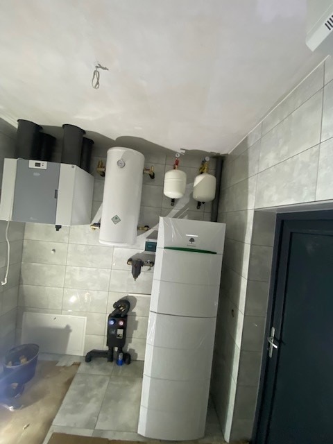 Nowo zainstalowana, owinięta folią pompa ciepła w kotłowni z szarymi płytkami, obok zbiornik buforowy i inne elementy instalacji grzewczej.