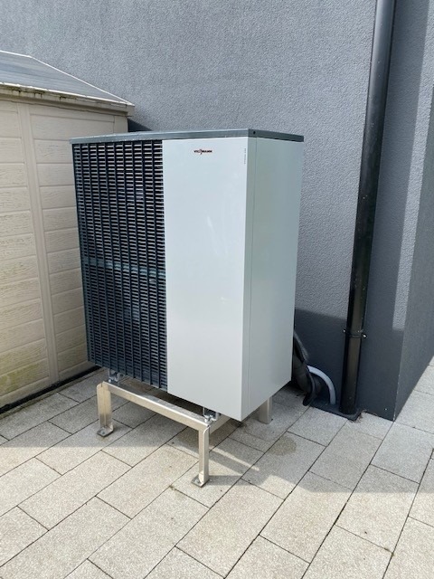 Zewnętrzna powietrzna pompa ciepła Viessmann na srebrnych nóżkach, ustawiona na szarej kostce brukowej przy ścianie budynku z rurą spustową.