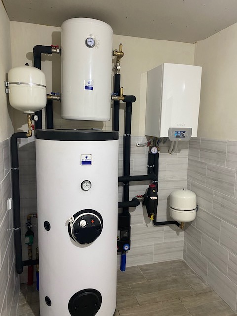 Zestaw urządzeń grzewczych w kotłowni: duży biały zasobnik ciepłej wody z czarnym panelem sterowania, mniejszy zbiornik wyrównawczy, wiszący kocioł gazowy i rury w izolacji, na tle szarych płytek...