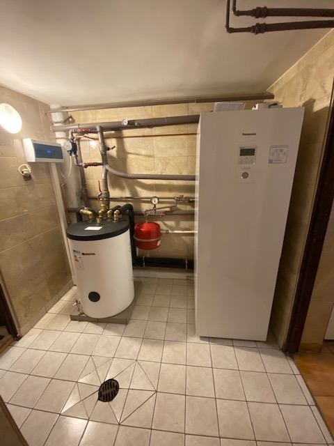 Biała pompa ciepła Panasonic obok cylindrycznego zbiornika na wodę w pomieszczeniu z beżowymi płytkami i kratką ściekową w podłodze.