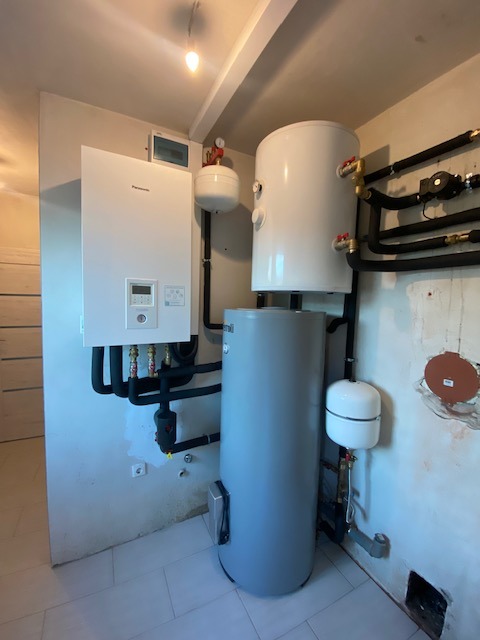 Zestaw kotła gazowego kondensacyjnego Panasonic, dwóch zbiorników akumulacyjnych i instalacji hydraulicznej w surowym pomieszczeniu z białymi płytkami podłogowymi.