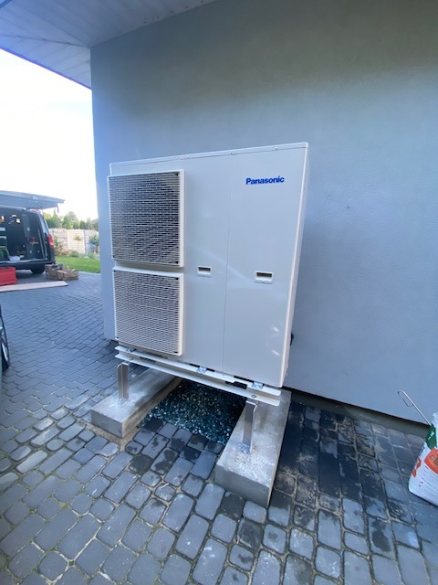 Zewnętrzna jednostka pompy ciepła Panasonic zamontowana na betonowych podstawach z metalowymi wspornikami, z widocznym drenażem wypełnionym drobnymi kamieniami, na tle szarej ściany budynku...