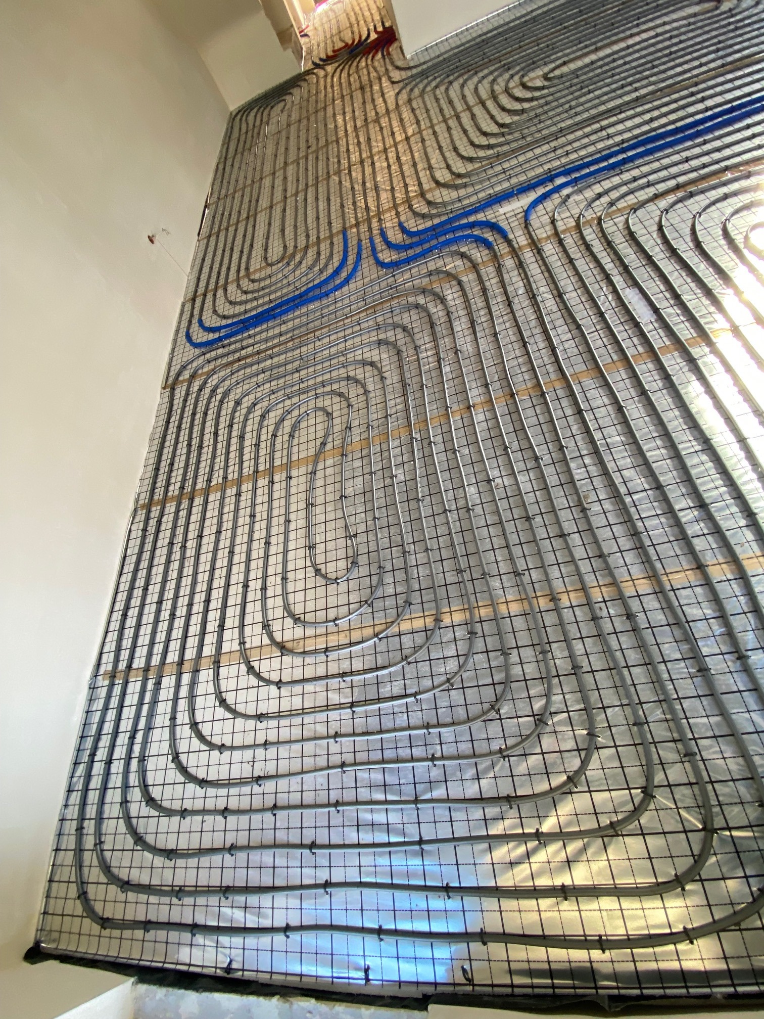 Szare rurki ogrzewania podłogowego ułożone w pętle na metalowej siatce z folią odblaskową, częściowo zalane betonem, widoczne niebieskie rury doprowadzające.