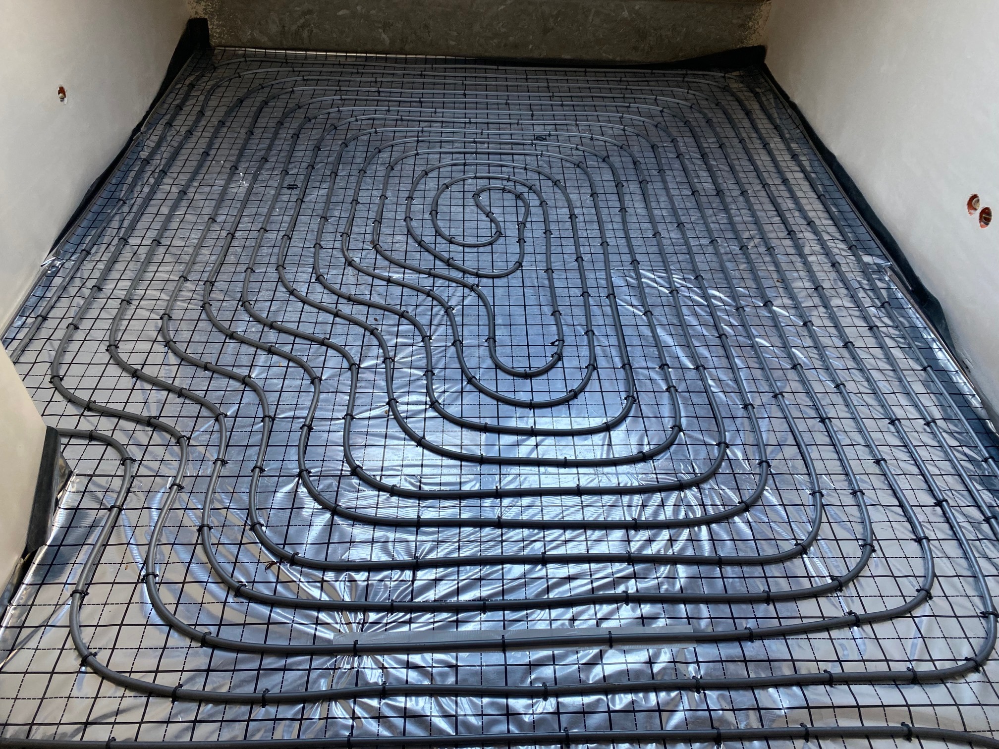 Instalacja ogrzewania podłogowego: szare rury ułożone spiralnie na srebrnej folii izolacyjnej z czarną siatką mocującą w surowym wnętrzu przed wylewką.