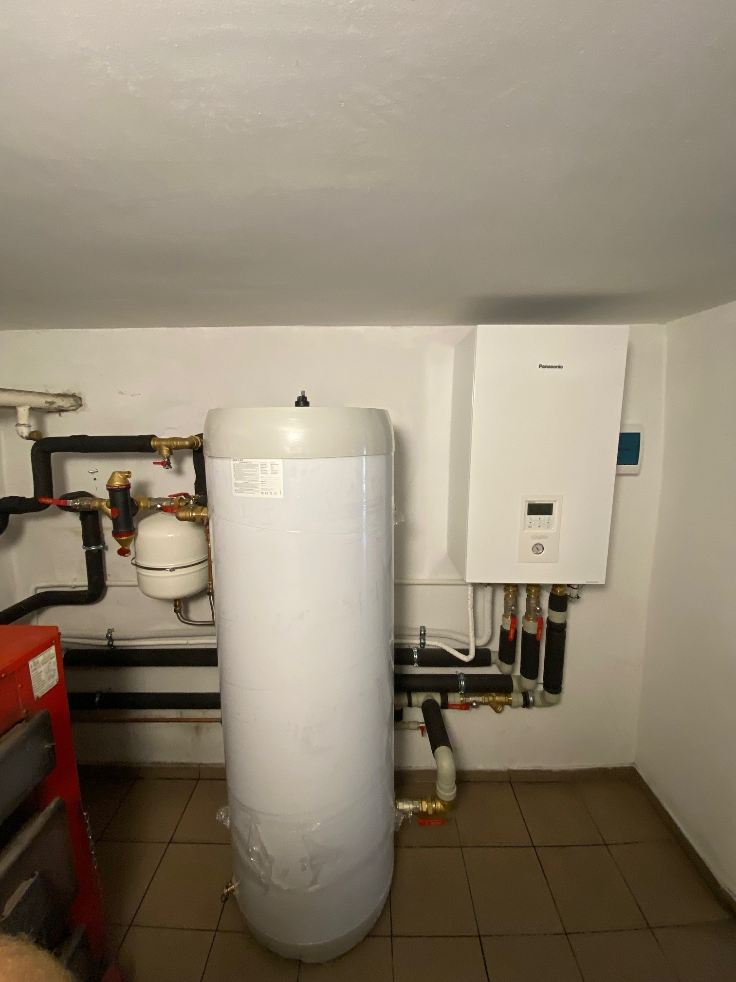Biały zasobnik ciepłej wody użytkowej, obok wiszący na ścianie biały moduł pompy ciepła Panasonic, widoczne rury i zawory, instalacja w pomieszczeniu technicznym.