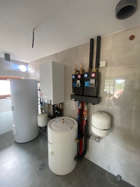 Kompletna instalacja grzewcza w kotłowni z dwoma zbiornikami akumulacyjnymi, kotłem, pompami i armaturą, rury w izolacji, szare płytki na ścianach i podłodze, widoczne okno.