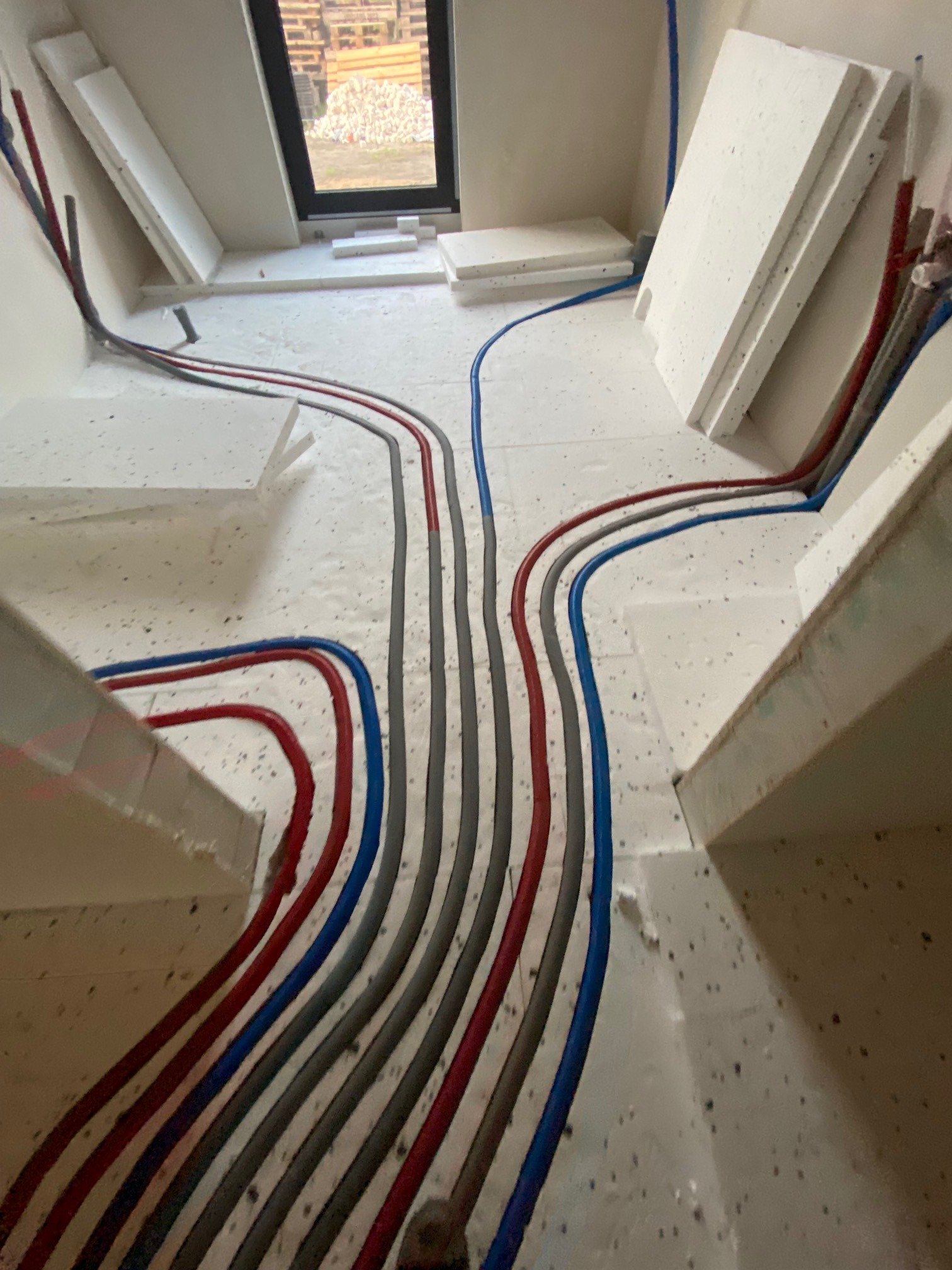 Wnętrze w trakcie instalacji ogrzewania podłogowego: widoczne rury w kolorach czerwonym, niebieskim i szarym ułożone na styropianie, przygotowane do zalania wylewką.