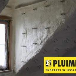 Poddasze po ociepleniu pianką PUR, widoczne metalowe uchwyty mocujące, okno w drewnianej ramie zabezpieczone folią oraz logo firmy izolacyjnej w prawym dolnym rogu.
