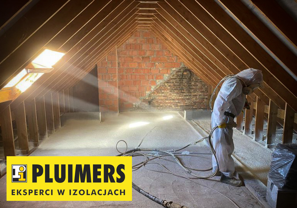 Pracownik w kombinezonie ochronnym aplikuje piankę izolacyjną na poddaszu z widoczną konstrukcją drewnianą i fragmentem ściany z cegły. W tle widoczne promienie światła wpadające przez okno dachowe...
