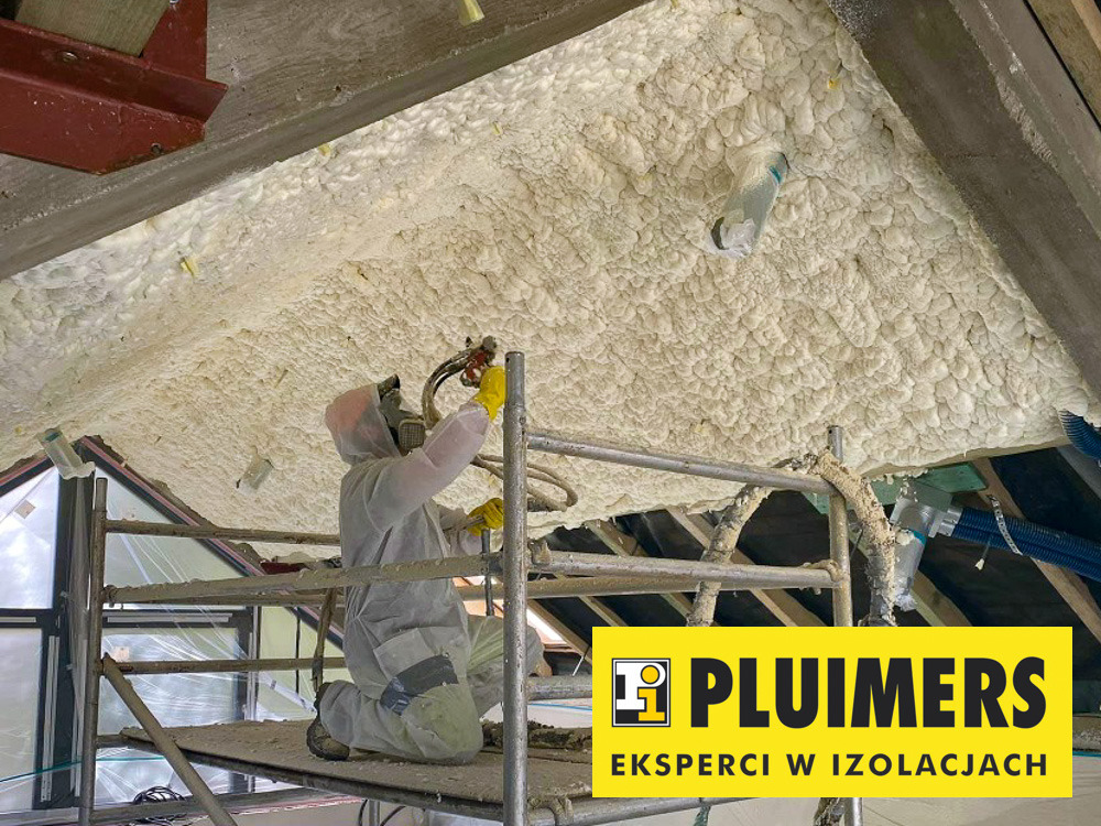 Pracownik w kombinezonie ochronnym aplikuje piankę PUR na poddasze, widoczne rusztowanie i zabezpieczone elementy konstrukcyjne.