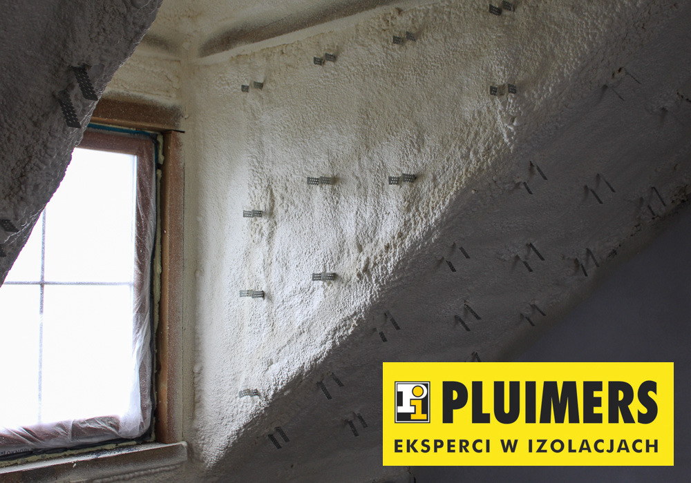Poddasze po ociepleniu pianką PUR, widoczne metalowe uchwyty mocujące, okno w drewnianej ramie zabezpieczone folią oraz logo firmy izolacyjnej w prawym dolnym rogu.