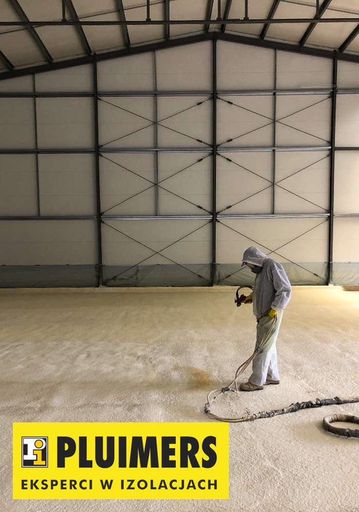 Pracownik w kombinezonie ochronnym natryskuje piankę izolacyjną na podłogę hali przemysłowej, widoczne elementy konstrukcji stalowej dachu.