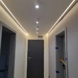 Nowoczesny korytarz z oświetleniem LED w suficie, punktowymi lampami i dwiema parami drzwi, z których jedne są zabezpieczone taśmą malarską i kawałkiem kartonu.