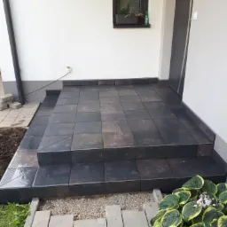 Wykończone schody wejściowe do domu z ciemnych, kwadratowych płytek, otoczone rabatami kwiatowymi i kamieniami ozdobnymi, widoczne gniazdko elektryczne na ścianie.