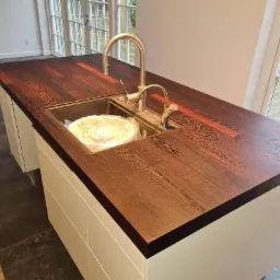 Wyspa kuchenna z blatem z ciemnego drewna z widocznym usłojeniem, z wbudowanym zlewem i baterią, białe szafki, okno w tle.