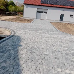 Ułożona szara kostka brukowa prowadząca do garażu z panelami słonecznymi na dachu, widoczne obrzeża i przygotowane podłoże pod trawnik.