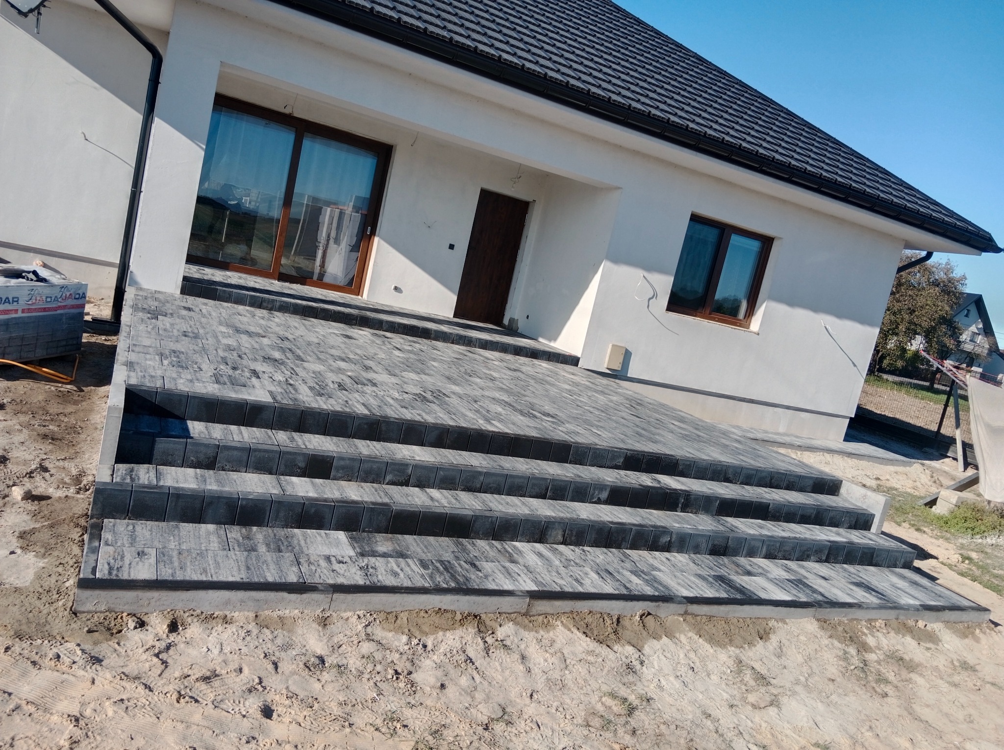 Schody wejściowe do domu jednorodzinnego wyłożone szarą kostką brukową, widok z dołu na elewację budynku z oknami i drzwiami wejściowymi, obok paczka z kostką.