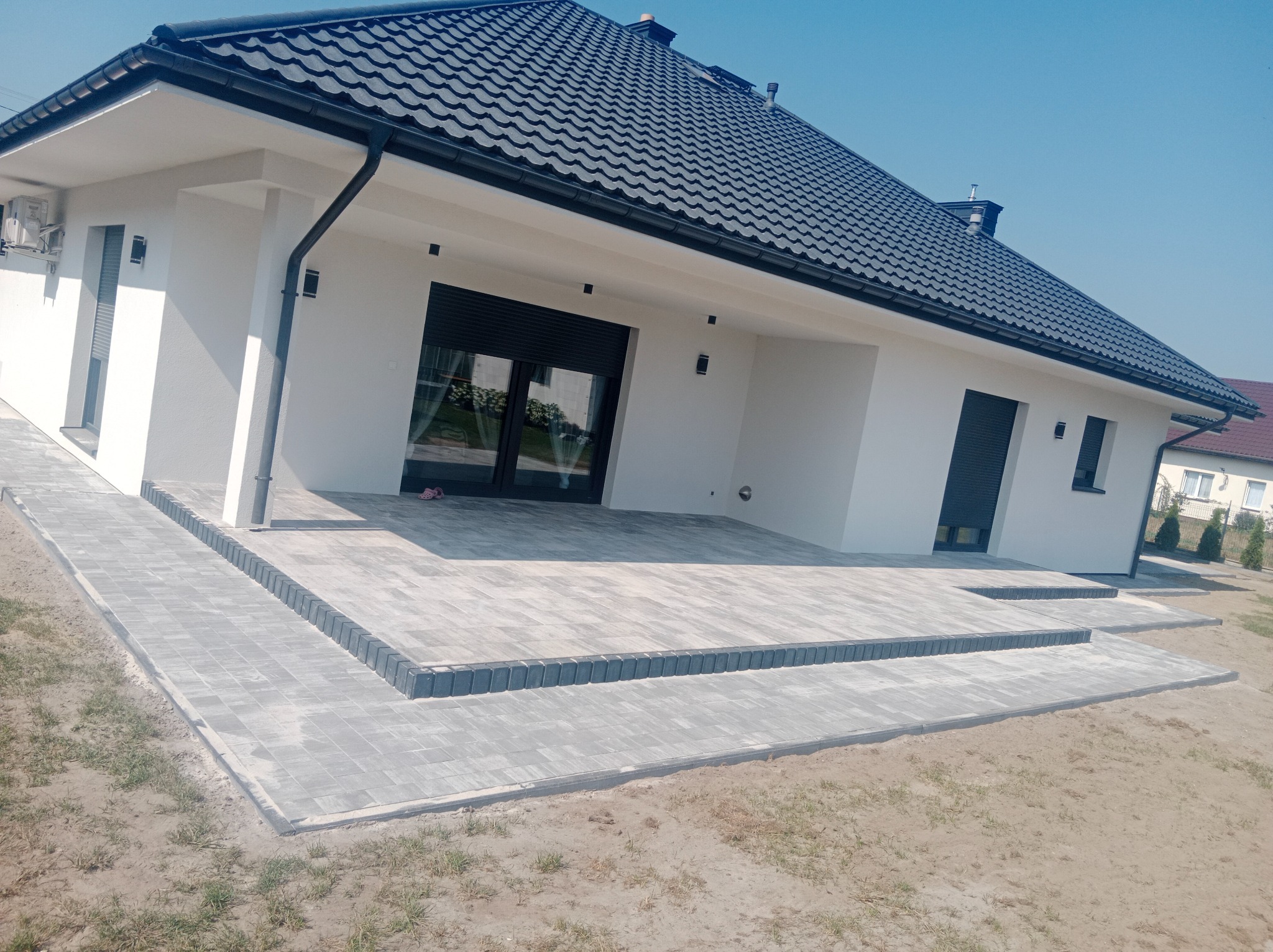 Nowoczesny dom z białym tynkiem i grafitowym dachem, z tarasem wyłożonym szarą kostką brukową z ciemnoszarym obramowaniem, widok z perspektywy trawnika.