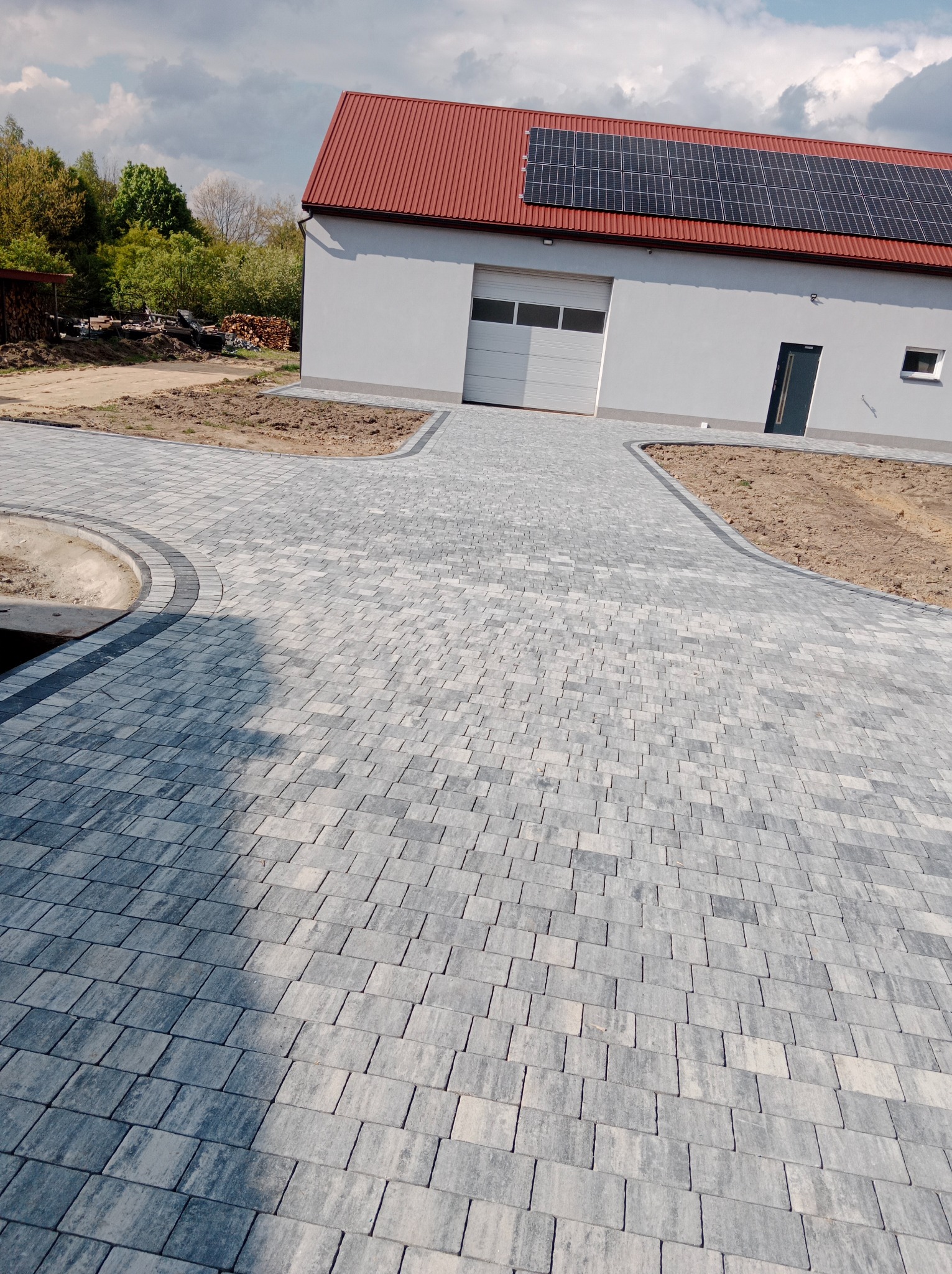 Ułożona szara kostka brukowa prowadząca do garażu z panelami słonecznymi na dachu, widoczne obrzeża i przygotowane podłoże pod trawnik.