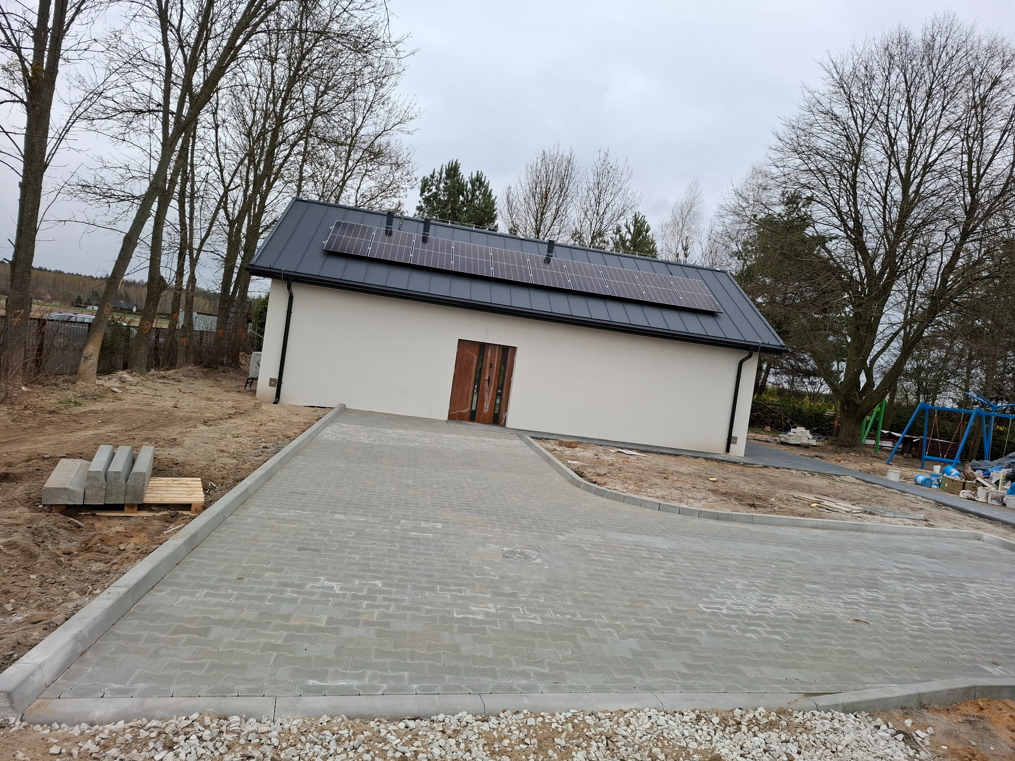 Nowo ułożona, szara kostka brukowa prowadząca do białego budynku z panelami słonecznymi na dachu, widoczne obrzeża i paleta z kostką w tle.