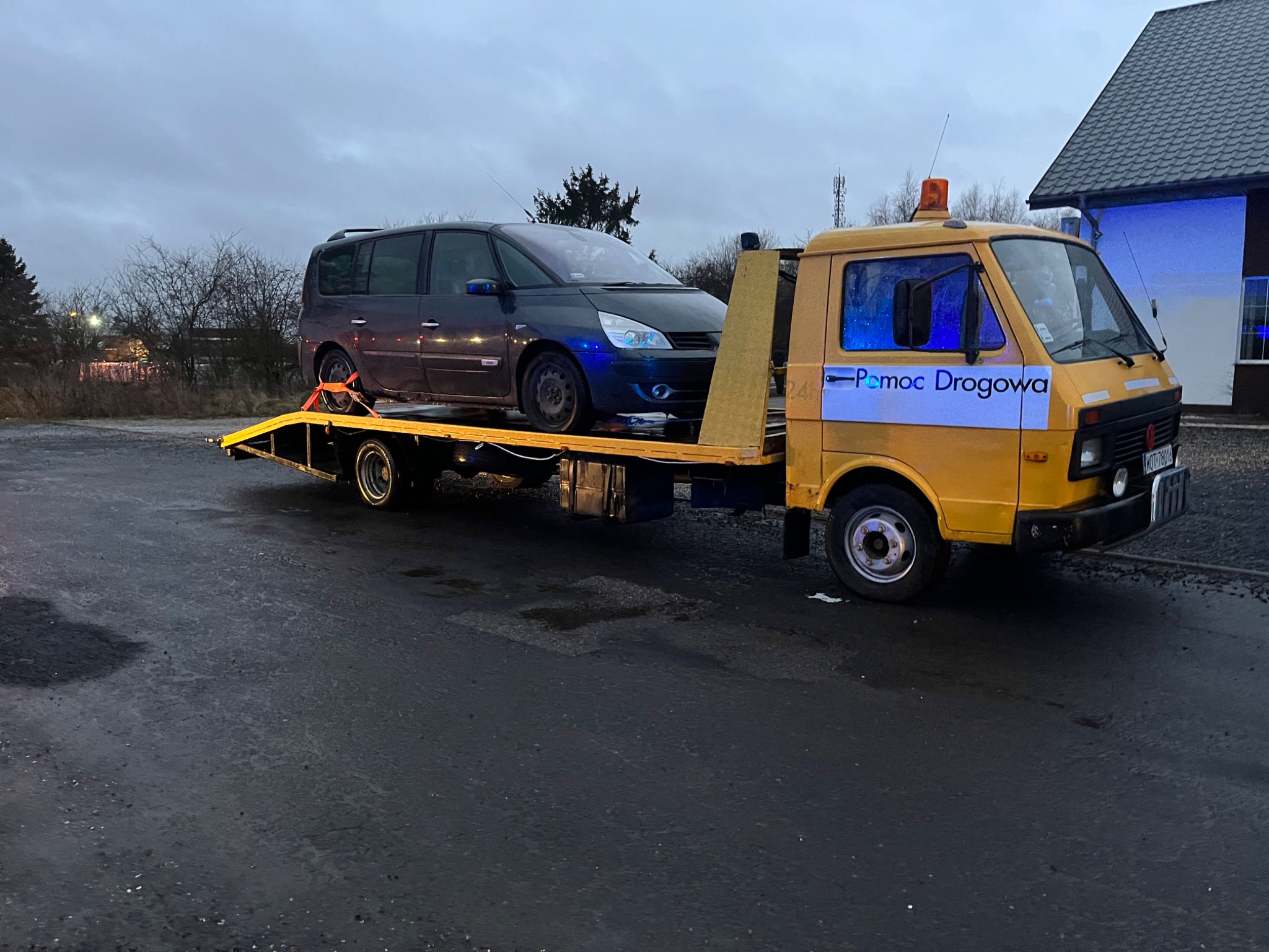 Żółta laweta z napisem 'Pomoc Drogowa' przewozi ciemny minivan na platformie, zabezpieczony pasami transportowymi, na tle szarego nieba i budynku z niebieskim oświetleniem.