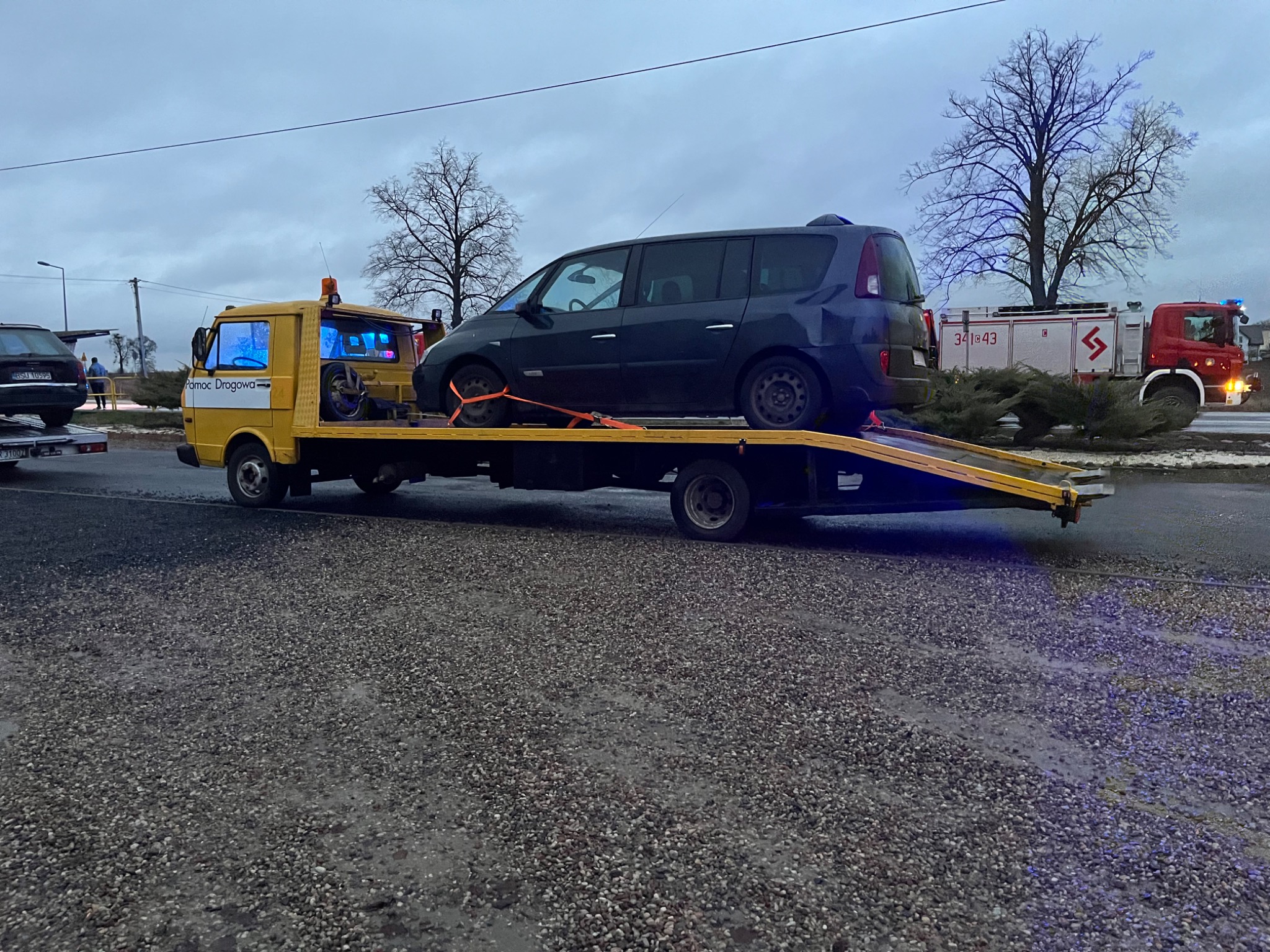 Żółta laweta z niebieskim minivanem zabezpieczonym pomarańczowymi pasami transportowymi, w tle wóz strażacki i drzewa bez liści, pochmurne niebo.