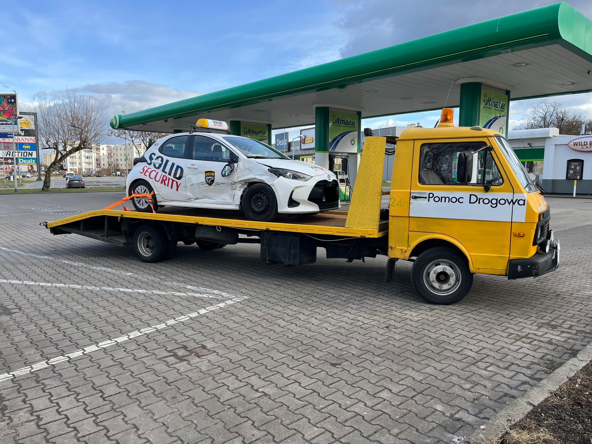 Żółta laweta z uszkodzonym białym samochodem osobowym firmy ochroniarskiej na platformie, zaparkowana na parkingu stacji benzynowej w pochmurny dzień.