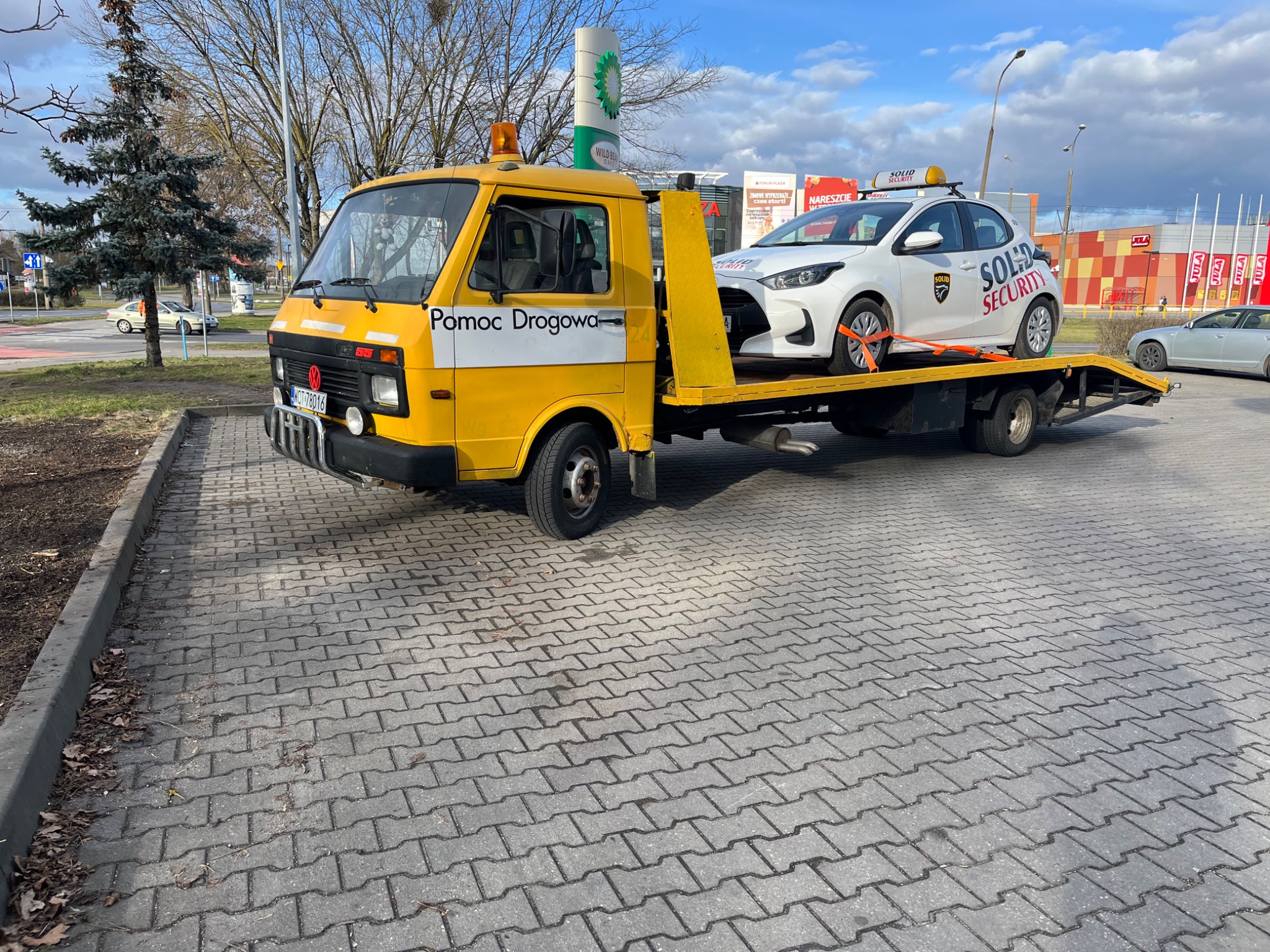 Żółta laweta 'Pomoc Drogowa' z białym samochodem 'Solid Security' na platformie, zaparkowana na parkingu przy sklepie, widoczny znak BP w tle.