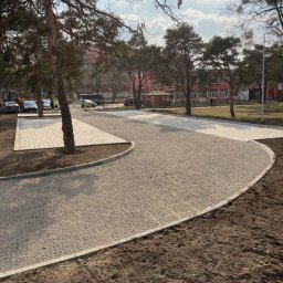 eR-BRUK Rafał Chojnacki - Nowo ułożona, jasnoszara nawierzchnia z kostki brukowej na podbudowie żwirowej, obrzeża z betonowych krawężników, w tle budynek i drzewa, prace w toku, widoczny sprzęt budowlany.