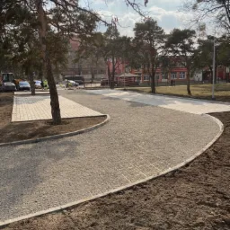 Nowo ułożona, jasnoszara nawierzchnia z kostki brukowej na podbudowie żwirowej, obrzeża z betonowych krawężników, w tle budynek i drzewa, prace w toku, widoczny sprzęt budowlany.