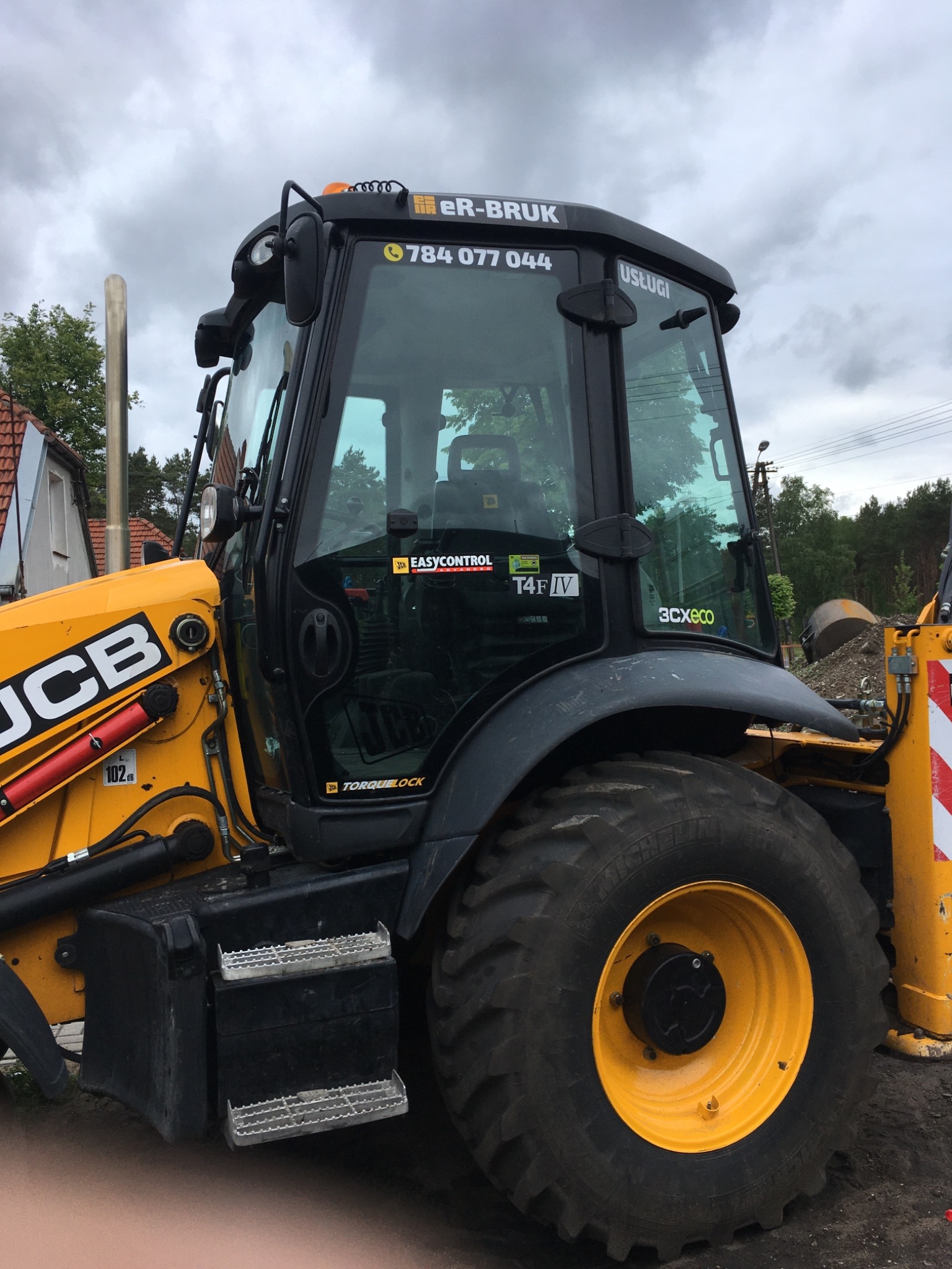 Żółto-czarna koparko-ładowarka JCB z widocznym numerem telefonu firmy 'eR-BRUK' na szybie kabiny, stojąca na ziemi.