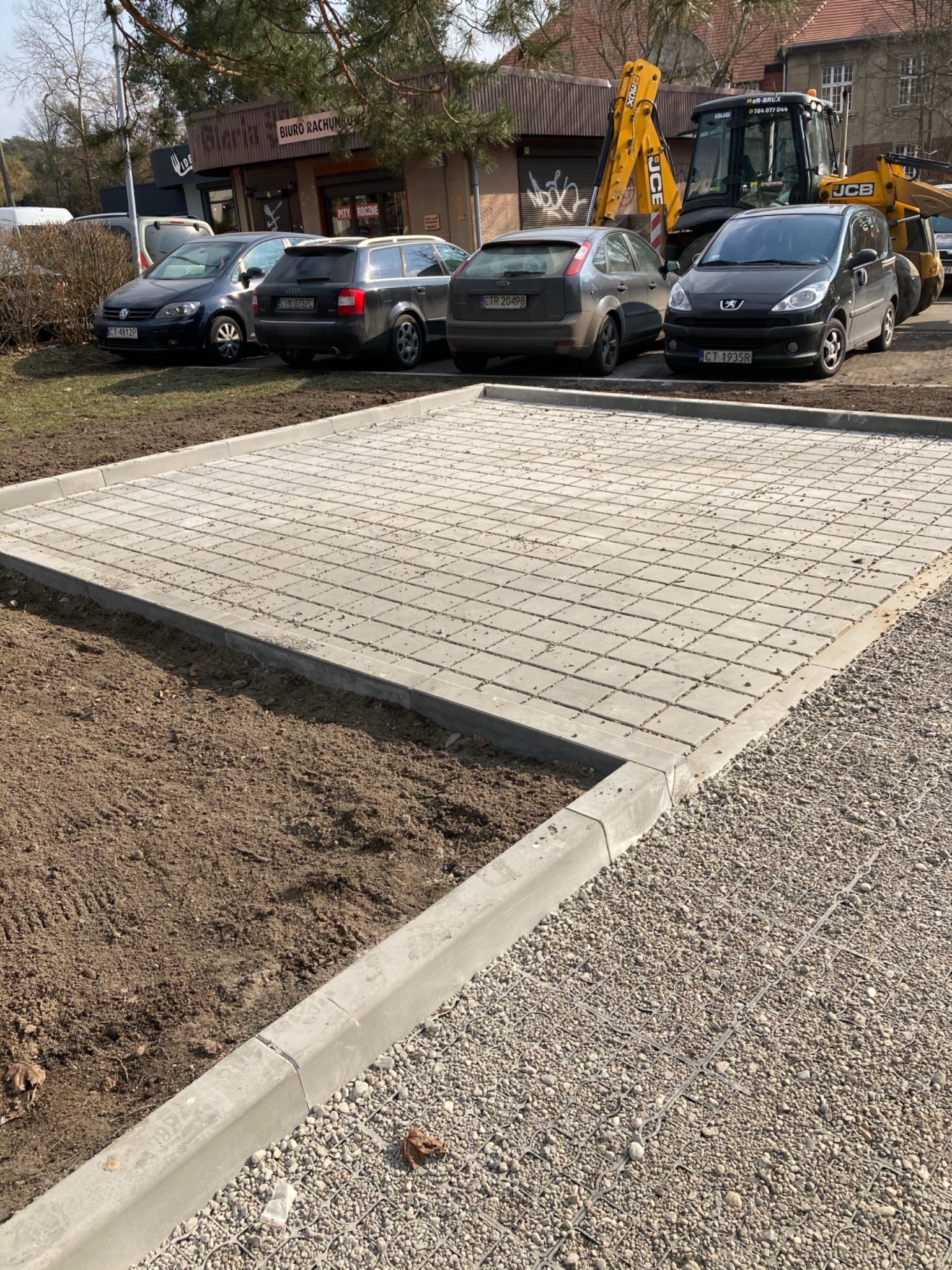 Ukończona powierzchnia z ażurowych płyt betonowych, obramowana krawężnikami, z przygotowanym podłożem z boku, na tle zaparkowanych samochodów i koparko-ładowarki JCB.