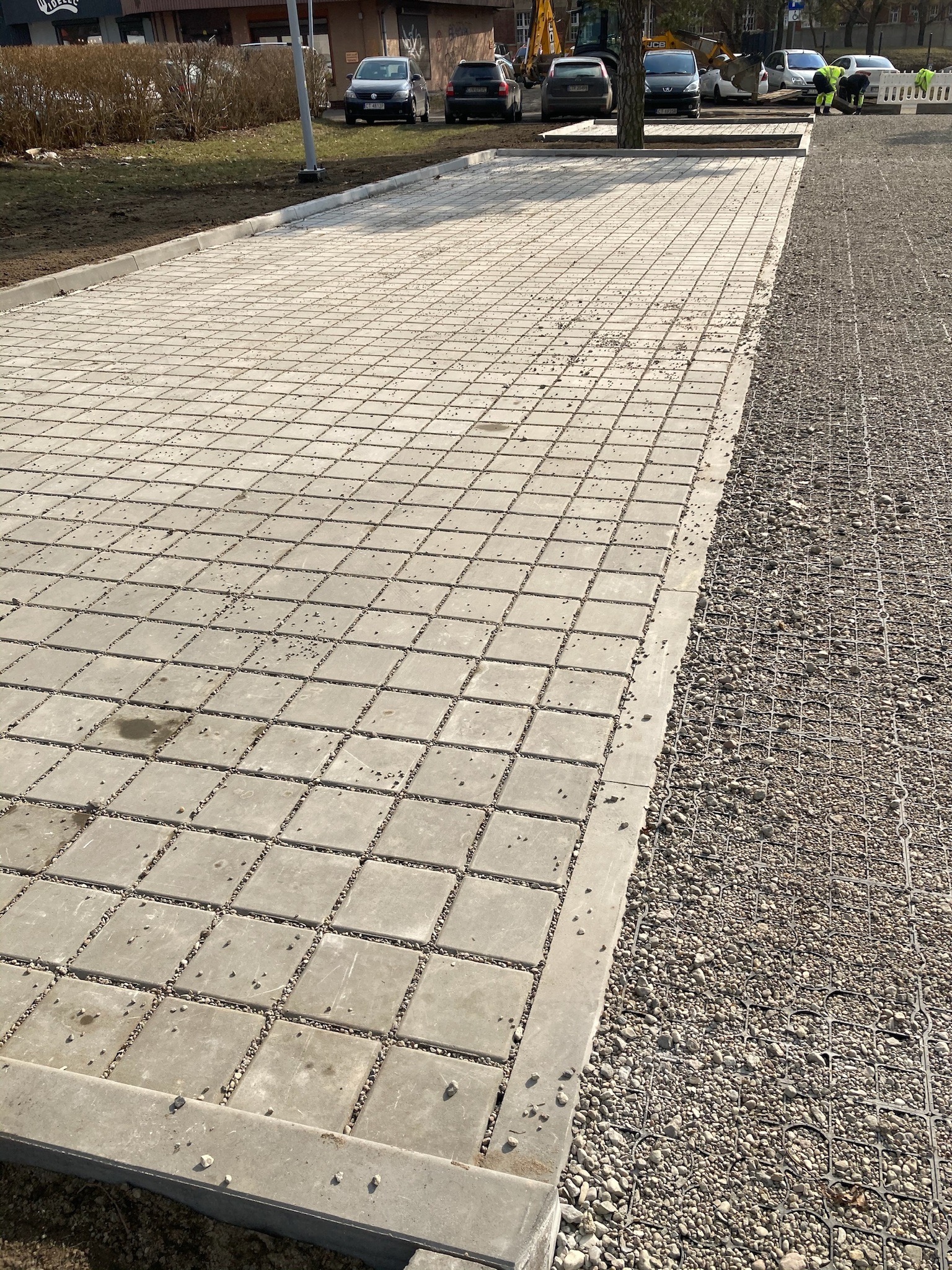 Ułożona nawierzchnia z szarej kostki betonowej na przygotowanym podłożu z kruszywa stabilizowanego siatką, widoczne krawężniki i pracownicy w tle.