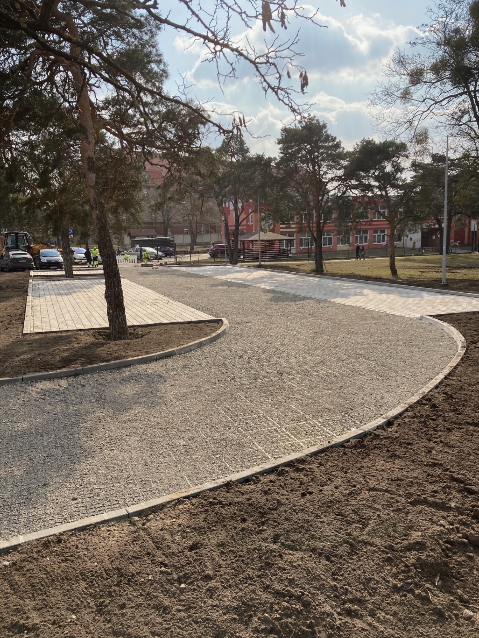 Nowo ułożona, jasnoszara nawierzchnia z kostki brukowej na podbudowie żwirowej, obrzeża z betonowych krawężników, w tle budynek i drzewa, prace w toku, widoczny sprzęt budowlany.